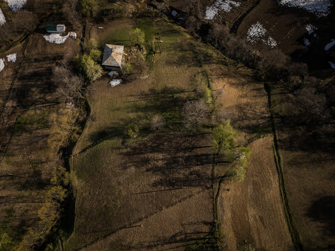 Aerial view of a quaint wooden house nestled amidst rolling hills, where patches of snow linger under the warm sunlight, Gorgadzeebi, Adjara, Georgia.