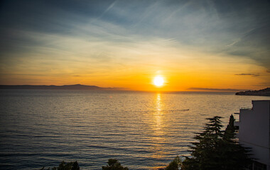 Stunning sunset over a serene beach in Croatia during the golden hour. The sky is painted in warm hues of orange, pink, and gold, casting a soft glow over the calm Adriatic Sea.