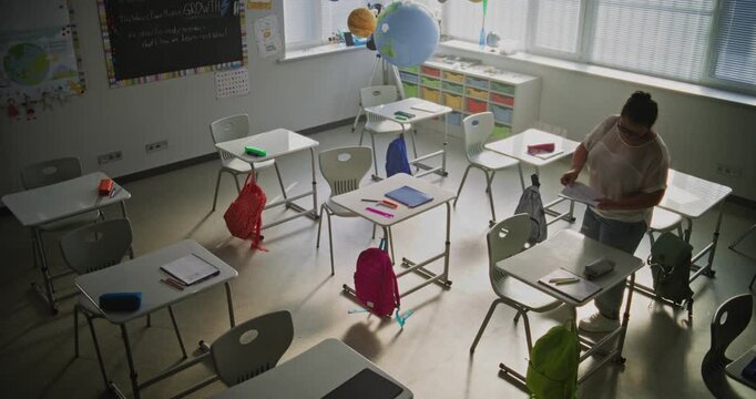 Female Teacher Laying Out Worksheets with Writing Exercises on Desks for School Children Before Lesson. Teacher Organizing Space for School Exam in Classroom, Working in Primary School. High Angle.