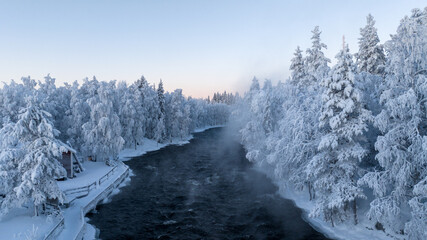 Aerial view of a dark, winding Kitkajoki river carving its path through a snow-laden forest, the trees draped in thick white blankets against the pale sky, Rovaniemi, Finland.