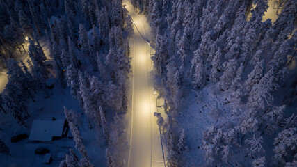 Aerial view of a ski trail through a dense, snow-laden forest, lights reflecting off the pristine white blanket, Salla, Lapland, Finland.