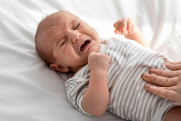 Closeup Portrait Of Crying Little Newborn Baby In Bodysuit Lying On Bed At Home, Upset Infant Child...