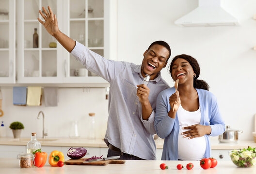 Joyful pregnant black couple having fun while cooking at home, happy african american expecting lady making dinner with her husband, singing songs, using spoons as microphones, kitchen interior