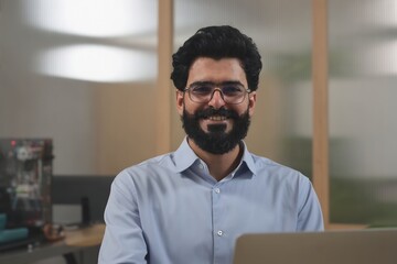 Portrait of a smiling man with beard and glasses working on laptop in office environment setting