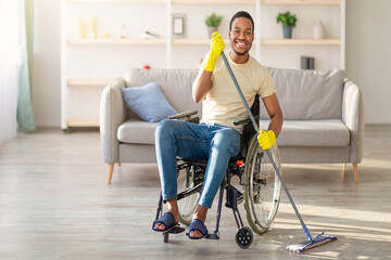Black man in wheelchair washing floor with mop at home. Happy guy with disability doing domestic...
