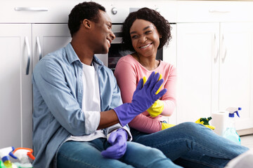 Smiling young black man and woman house-keepers sitting on floor in kitchen and giving each other...
