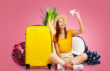A young woman with a big smile is sitting beside a bright yellow suitcase, surrounded by colorful...