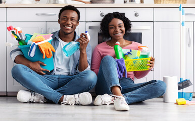 Cheerful black couple in rubber gloves sitting on floor next to oven, holding house-keeping...
