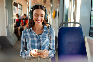 Happy lady sitting in modern tram, wearing wireless headphones and using cellphone, listening music...