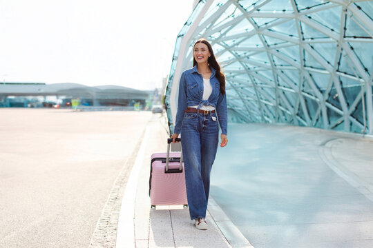 Young lady tourist with suitcase walking outdoor at modern airport terminal or train station, waiting for flight boarding, ready for vacation, full length