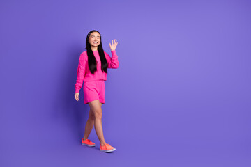 Young woman in stylish pink outfit walking gracefully against a vibrant purple background, waving...