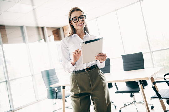 Confident young businesswoman working on a tablet in a professional office workspace with large windows and modern furniture.