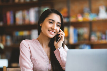 Excited latin woman entrepreneur talking on smartphone while working on laptop computer in cafe,...
