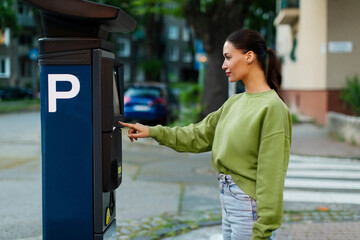 Young woman paying for parking, using outdoors electronic pay station terminal, lady driver using...