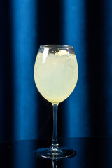 Refreshing light yellow cocktail served in a wine glass with ice and a lemon wedge. The drink is set against a deep blue background, creating a cool, summery, and elegant visual impression