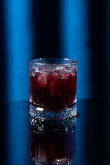 Close-up of a dark red cocktail with ice cubes served in a faceted old fashioned glass. The drink is rich in color and presented against a deep blue background, creating a bold and moody visual impres