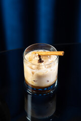 Creamy iced coffee cocktail in a short glass with a cinnamon stick and coffee beans on top, served on a glossy black surface with a dark blue background
