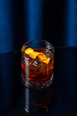 Close-up of a Negroni cocktail with ice and an orange peel garnish in a glass on a dark glossy surface, with a deep blue background. Stylish and moody bar setting