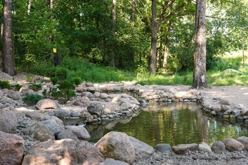  A tranquil park setting features a small waterfall cascading over rocks into a serene pond. The peaceful scene is enhanced by a person exploring the lush, wooded surroundings.