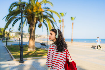 Carefree student enjoying music while walking on a seaside promenade with palm trees, carrying a...