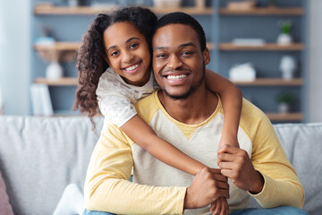 Closeup portrait of happy black father and daughter embracing while having fun together at home, copy space. Smiling african american school girl hugging her cheerful dad from behind