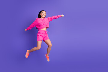 Happy young woman in bright pink clothing jumping joyfully against a vivid purple background, full...