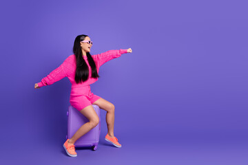 Fashionable young woman in vibrant pink outfit posing playfully with purple suitcase against...