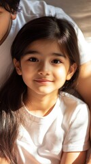 A young girl with long hair is smiling at the camera. She is wearing a white shirt and is sitting on a couch