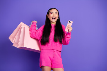 Cheerful young woman with shopping bags wearing pink outfit holding smartphone against violet to...