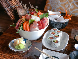 Bowl of assorted sashimi with salmon rolls, sweet potato fries, and dipping sauce on wooden table.