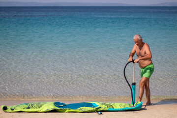 A mature man inflating his paddle board on the beach with a sea background. A senior male getting ready for some fun on a sunny day.