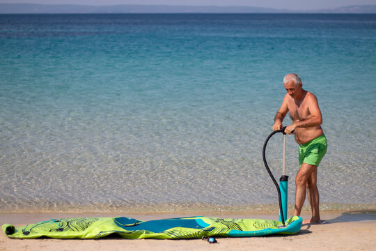 A mature man inflating his paddle board on the beach with a sea background. A senior male getting ready for some fun on a sunny day.