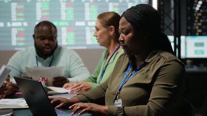 African american analyst and colleagues examining annual data reporting in a firm meeting at night, reviewing insight on laptop to facilitate global business development. Work team. Camera B. - Powered by Adobe