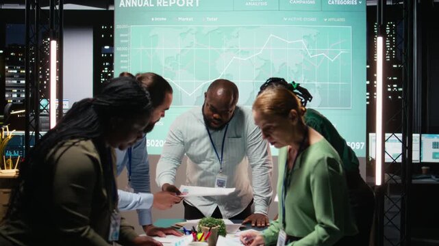 Diverse coworkers team participating in a late night briefing session, exchanging documents and reviewing finance updates. Aligning objectives through collaborative problem solving. Camera A.