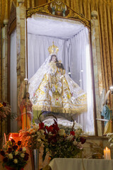 Virgin of the Assumption on an altar, illuminated by white light, surrounded by floral decorations, candles, and two angels. Located in Huata, province of Huaylas, Ancash - Peru