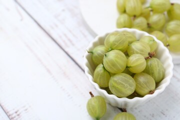 Fresh green gooseberries in bowl on white wooden table, closeup. Space for text