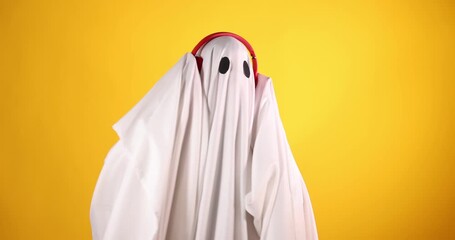 Funny ghost. Man covered with white sheet listening to music with headphones on orange background