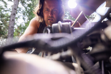Man inspects a car engine using a flashlight in an outdoor wooded setting. Hands-on automotive...