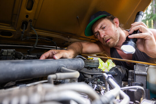 Close-up of a man wearing a cap using a flashlight to inspect a car engine. Detailed view of vehicle maintenance and mechanical work under the hood.