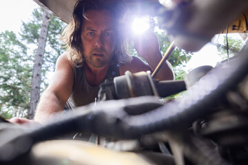 Man inspects a car engine using a flashlight in an outdoor wooded setting. Hands-on automotive...