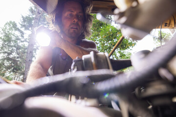 Man inspects a car engine using a flashlight in an outdoor wooded setting. Hands-on automotive...