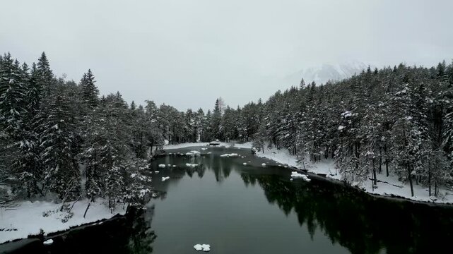 Stunning drone pull back from a tranquil winter lake to reveal a picturesque village in a vast, snow-covered alpine valley with mountains.
