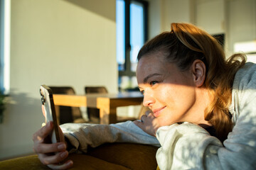Relaxed woman smiling while scrolling on smartphone in cozy sunlit living room