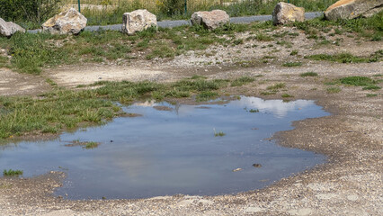 A large muddy puddle reflecting the cloudy sky on a dirt and gravel path after rain