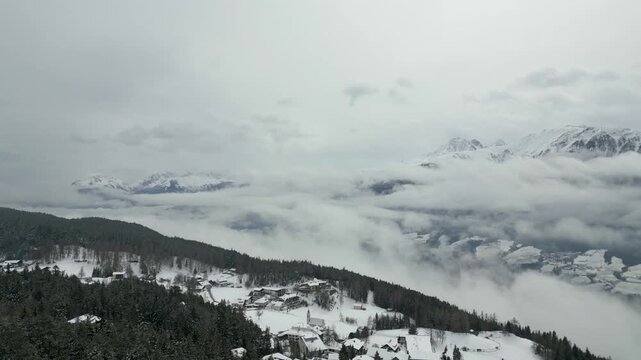 Winter Alpine Village: An aerial view of a picturesque snow-covered town nestled on a forested mountainside, with majestic peaks emerging from swirling clouds.