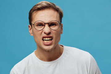 Frustrated young man with glasses expressing anger and annoyance against bright blue background,...