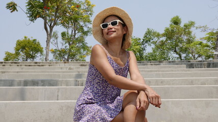 An Asian female model in a floral dress and a straw hat sits with her legs crossed on concrete...