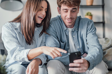 Happy millennial couple sit relax on couch in living room watching video on cellphone together,...