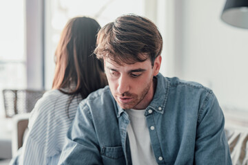 Attractive man is depressed, sitting on the couch ignoring woman, having a dispute with wife or...