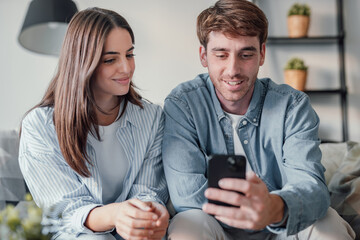 Happy millennial couple sit relax on couch in living room watching video on cellphone together,...